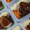 Plastic meal prep containers filled with roast beef covered in gravy, mashed potatoes, peas, and roasted carrots. The dishes appear hearty and comforting.