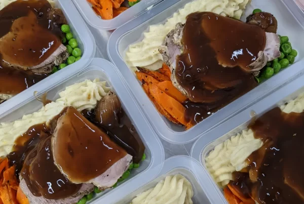 Plastic meal prep containers filled with roast beef covered in gravy, mashed potatoes, peas, and roasted carrots. The dishes appear hearty and comforting.