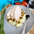 A plate with Three slices of poached chicken breast on top of a creamy potato salad with crispy bacon and cucumber pickle.