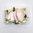 Three slices of poached chicken breast on top of a creamy potato salad with crispy bacon and cucumber pickle in a meal tray.