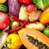 A colourful assortment of fresh fruits and vegetables including carrots, lettuce, broccoli, papaya, pomegranate, corn, tomatoes, capsicum, garlic, and eggplant displayed on a wooden table.