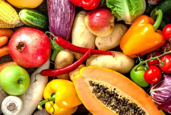 A colourful assortment of fresh fruits and vegetables including carrots, lettuce, broccoli, papaya, pomegranate, corn, tomatoes, capsicum, garlic, and eggplant displayed on a wooden table.