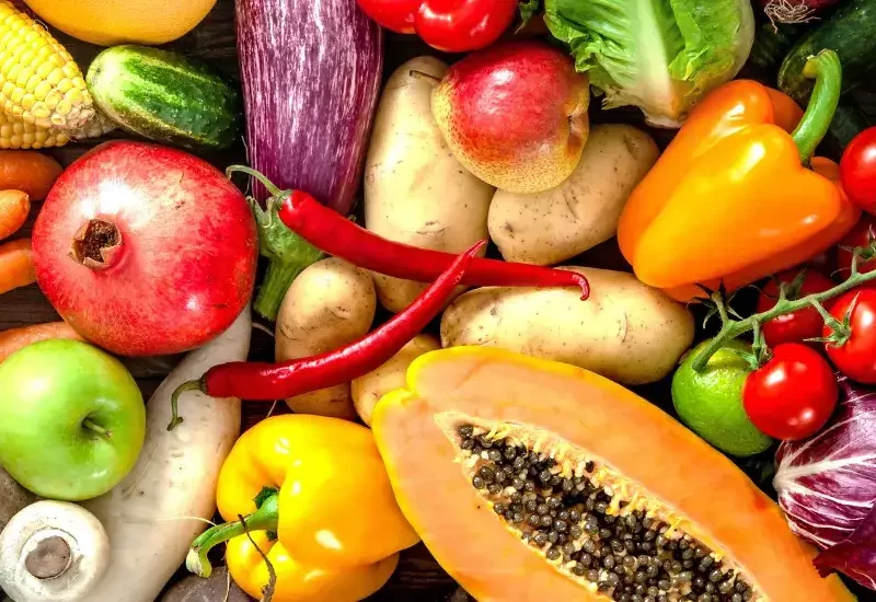 A colourful assortment of fresh fruits and vegetables including carrots, lettuce, broccoli, papaya, pomegranate, corn, tomatoes, capsicum, garlic, and eggplant displayed on a wooden table.